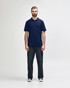 A man stands wearing a navy blue Gildan Softstyle adult double pique polo and black jeans. The polo features a logo.