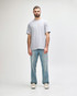 A man stands wearing a light grey Gildan Softstyle adult midweight T-shirt and blue jeans. The shirt has a logo.