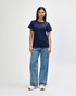 A woman wearing a navy Gildan Softstyle ladies midweight T-shirt, paired with blue jeans and white sneakers.