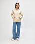 A hooded sweatshirt in cream worn by a person, paired with blue jeans and white shoes. The sweatshirt has a logo.