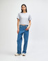 A woman wearing a grey boxy t-shirt and blue jeans, standing against a plain background. The shirt has a logo.