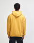 A unisex hooded sweatshirt in mustard yellow, featuring a spacious fit and a hood at the back. It has a branding logo.