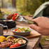 A fork with a green handle holds grilled sausage and vegetables, with camping gear and food in the background.