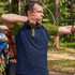 A man in a navy and green polo shirt is aiming an arrow with a bow in a forest setting. The shirt has a logo.