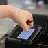 A hand wearing a dark ceramic ring taps a contactless payment terminal. The terminal has a display screen.