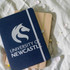 A navy A5 notebook with a logo, placed atop a beige notebook, surrounded by flower petals on a soft surface.