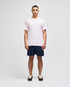 A man stands wearing a light pink short sleeve t-shirt and navy shorts, featuring a logo on the t-shirt.