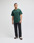 A green short sleeve t-shirt worn by a standing man, paired with dark pants and shoes. The shirt has a logo.