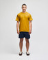 A mustard yellow short sleeve t-shirt displayed on a model, paired with navy shorts. The shirt features a logo.