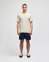 A beige short sleeve t-shirt displayed on a male model, paired with navy shorts. The shirt features a logo on the sleeve.