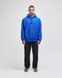 A blue pullover hooded sweatshirt with a front pocket, worn by a standing model in casual black pants.
