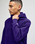 A purple hooded sweatshirt with a logo on the sleeve, worn by a model. The sweatshirt features a high neckline.