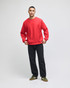 A red adult crew sweatshirt worn by a model, paired with black pants and green sneakers. The sweatshirt has a logo.