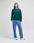 A green crewneck sweatshirt is worn with light blue jeans by a standing model in a neutral background. The sweatshirt has a logo.
