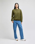 An olive green sweater worn by a model with blue jeans and white shoes, standing against a light background.