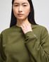 An olive green crewneck sweatshirt featuring a logo on the sleeve, modelled by a woman.