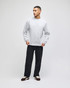 A grey adult crewneck sweatshirt displayed on a model, paired with black trousers and shoes. The sweatshirt features a logo.