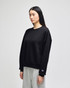 A black crewneck sweatshirt is displayed on a model, featuring a logo on the sleeve. The model wears grey pants.