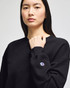 A black women's crewneck sweatshirt with a logo on the sleeve. The model is posing with a serious expression.