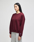 A maroon women's crewneck sweatshirt with a relaxed fit, featuring a logo on the sleeve. Styled with grey pants.