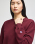 A burgundy women's crewneck sweatshirt featuring a logo on the sleeve. The model is standing and looking forward.