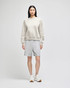 A model wearing a light grey crewneck sweatshirt and grey shorts, paired with white sneakers. The sweatshirt has a logo.