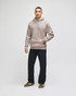 A man stands wearing a beige hooded sweatshirt and dark jeans, with sneakers. The sweatshirt has a logo.