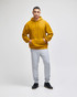 A mustard yellow pullover hooded sweatshirt worn by a model with grey track pants and black shoes. The sweatshirt has a logo.