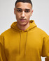 A mustard yellow pullover hooded sweatshirt with a drawstring hood, worn by a model. It features a logo on the garment.