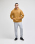 A hooded sweatshirt in a warm mustard colour paired with light grey sweatpants. The model is standing confidently.