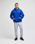 A blue pullover hooded sweatshirt with a front pocket, paired with grey sweatpants. The model stands confidently.