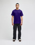 A purple short-sleeve t-shirt worn by a model, paired with black pants. The shirt features a logo.