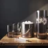 A glass carafe with ice in a metal bucket, accompanied by two clear glasses, all on a wooden surface.