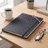 A black A4 diary laid on a wooden desk, accompanied by a pen, glasses, a coffee cup, and a small plant.
