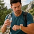 A man in a blue shirt holds a water bottle while looking at a green and pink Medicine Box Wristband on his wrist.