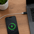 A USB data blocker in black connected between a phone and a laptop on a wooden surface, with a potted plant nearby.