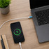 A smartphone charging beside a laptop, connected by a white USB cable on a wooden desk, with a green charging indicator.