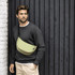 A waist bag in light green worn across a man's chest, with a casual grey sweater and beige pants, against a dark wooden background.