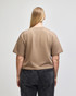 A back view of a beige mid-length boxy top with short sleeves, paired with dark grey pants. The top features a logo.