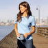 A woman in a light blue polo shirt stands on a pier, holding a drink can, with a city skyline in the background.