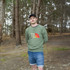A man stands in a forest wearing a green unisex sweatshirt with a logo. He pairs it with denim shorts.