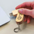 A wooden 8GB flash drive with a keychain attachment, held in a person's hand, next to a laptop.