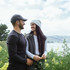 A woman wearing a grey vest and beanie stands beside a man, both smiling, in a scenic outdoor setting with flowers.