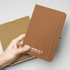A hand holding a brown hardcover notebook with an elastic band, featuring a logo on the front.