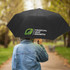 A black umbrella with a logo, held by a person wearing a denim jacket, on a tree-lined path.