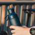 A drink bottle in dark blue, with a handle and a logo, held in a hand near a bike.