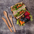 A lunch box filled with wholemeal wraps, strawberries, blueberries, celery sticks, and almonds, accompanied by wooden utensils.