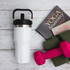 A drink bottle in white with a black lid and handle, beside a yoga book, mat, and pink dumbbells.