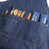 A denim blue bib apron featuring multiple pockets with wooden utensils and cloth napkins neatly arranged.