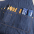 A poly-cotton denim full bib apron in navy, featuring pockets filled with utensils and two rolled towels.